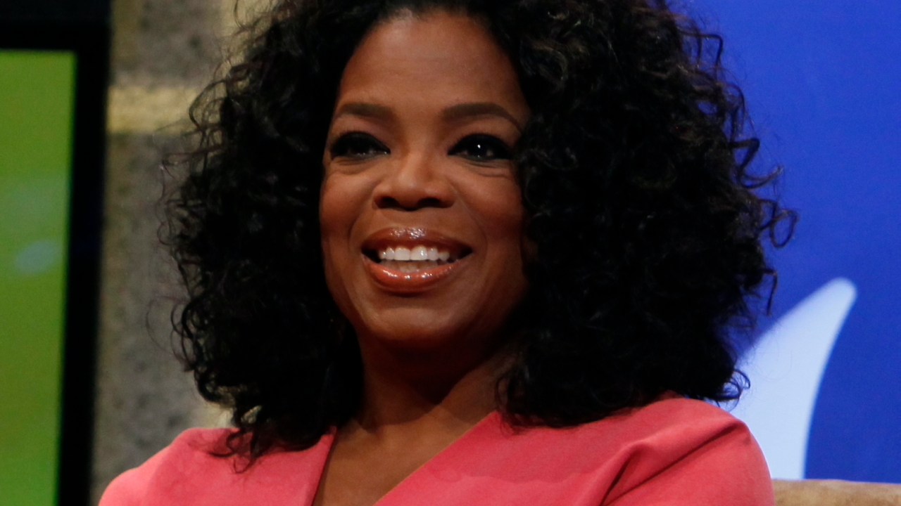 Mulher negra, de cabelo afro, sorri, usando um blazer rosa. A foto é do busto para cima.