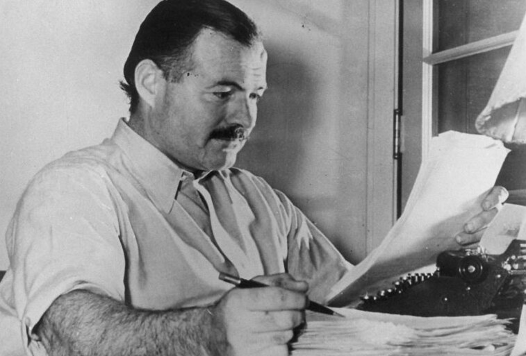 O escritor Ernest Hemingway. Um homem branco, de meia idade, cabelos negros e bigode. Ele está sentado em uma mesa, com um lápis na mão e um papel na outra. Ele olha para o papel