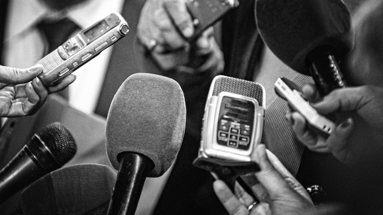 Imagem em preto e branco mostra vários microfones estendidos como em uma entrevista coletiva