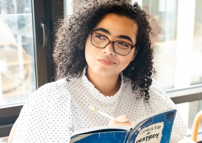 Garota negra pensativa, com livro e caneta na mão.