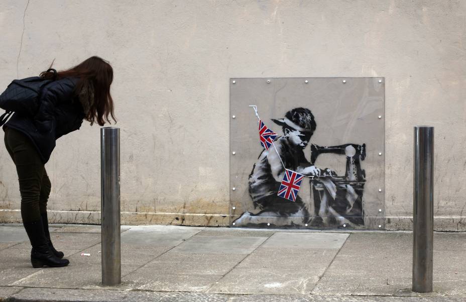 <span>Neste outro grafite do artista britânico Banksy, a imagem nos mostra uma criança em uma máquina de costura, produzindo bandeiras do Reino Unido. A pintura foi feita na parede de uma loja de artigos populares, no norte de Londres. Muitas confecções em países em desenvolvimento, principalmente do sudeste asiático, exploram o trabalho infantil e vendem seus produtos para famosas redes varejistas nas nações mais ricas. O grafite de Banksy é uma clara crítica a esta situação.</span> (foto: Peter Macdiarmid/Getty Images)