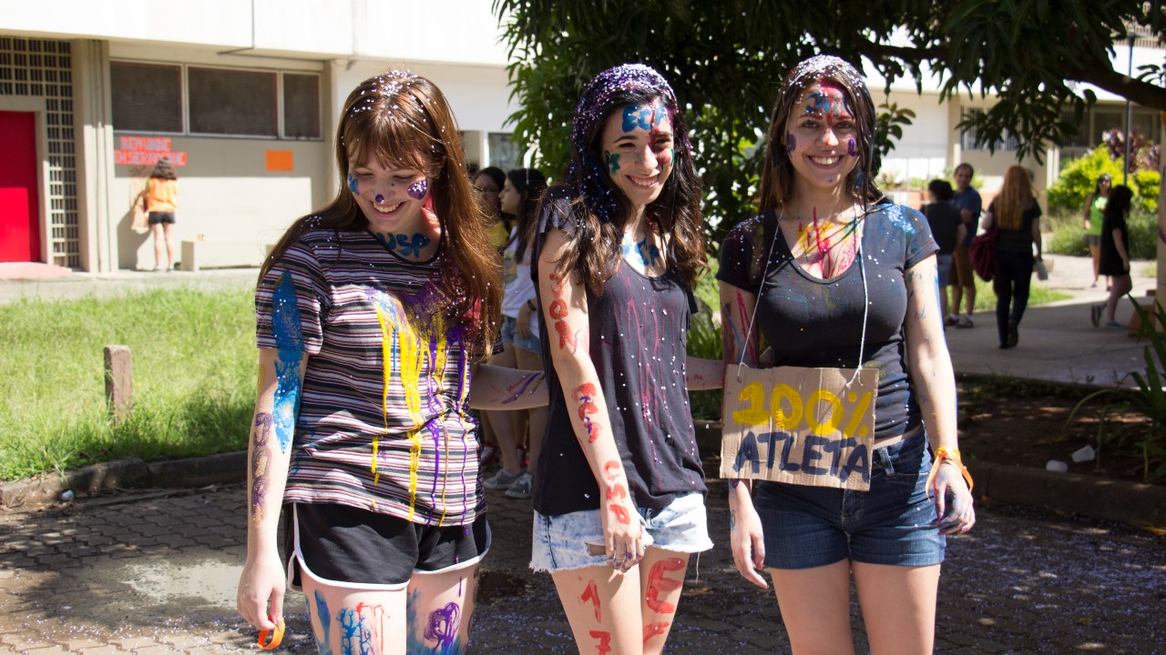 Três meninas sorriem e posam cobertas de tinta nos cabelos, com uma segurando uma placa escrito "100% atleta". As três estão no campus da Universidade de São Paulo.