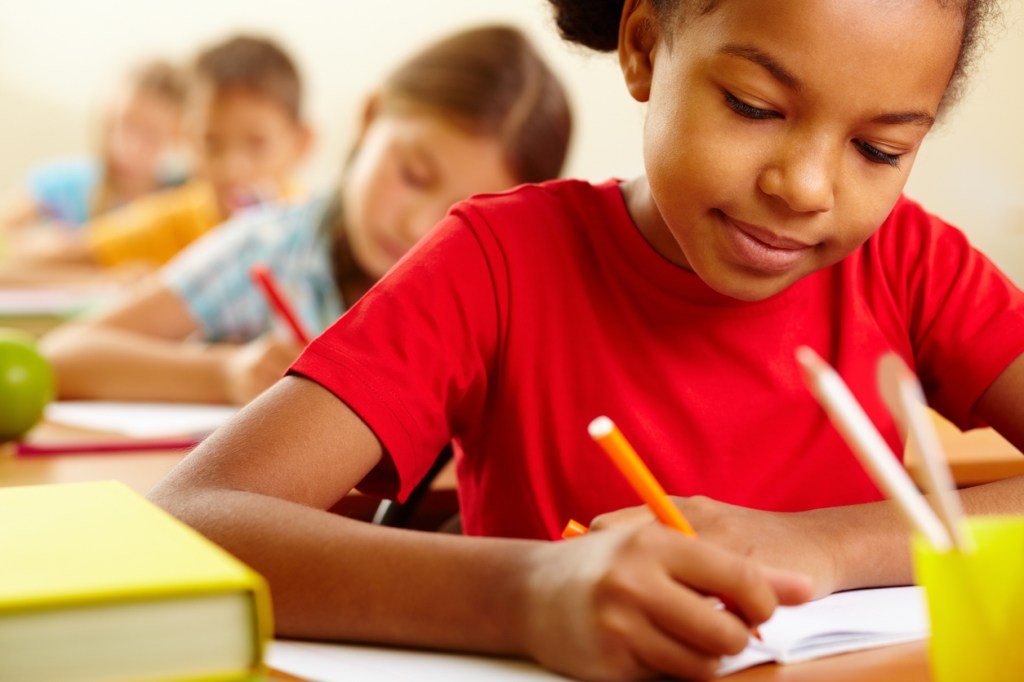 Crianças escrevendo em sala de aula. A fotografia foca em uma garota negra, segurando um lápis na mão e com o semblante concentrado.