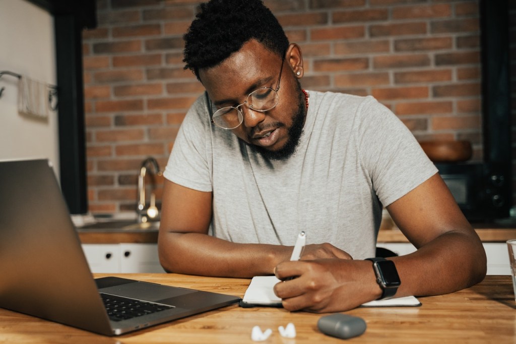Homem negro sentado escrevendo com um notebook aberto de frente para ele.