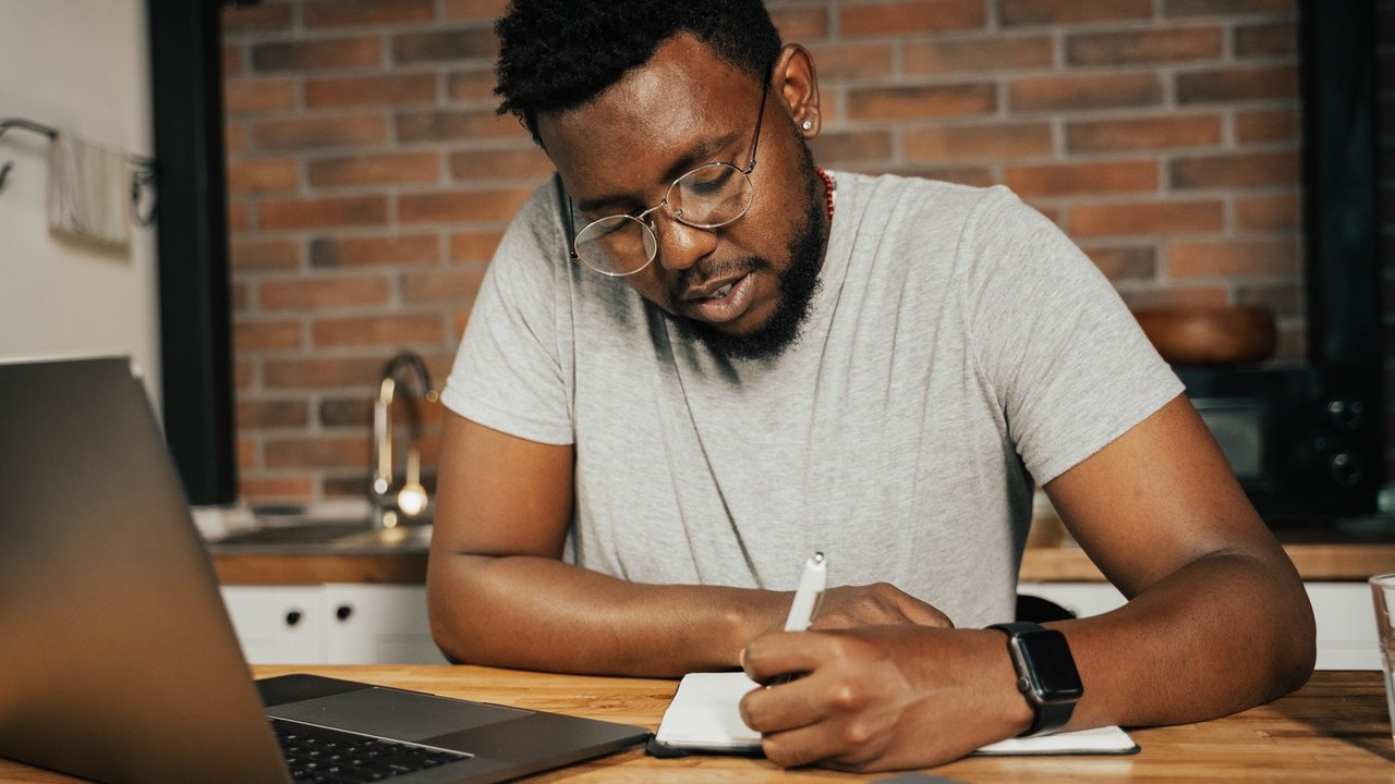 Homem negro sentado escrevendo com um notebook aberto de frente para ele.