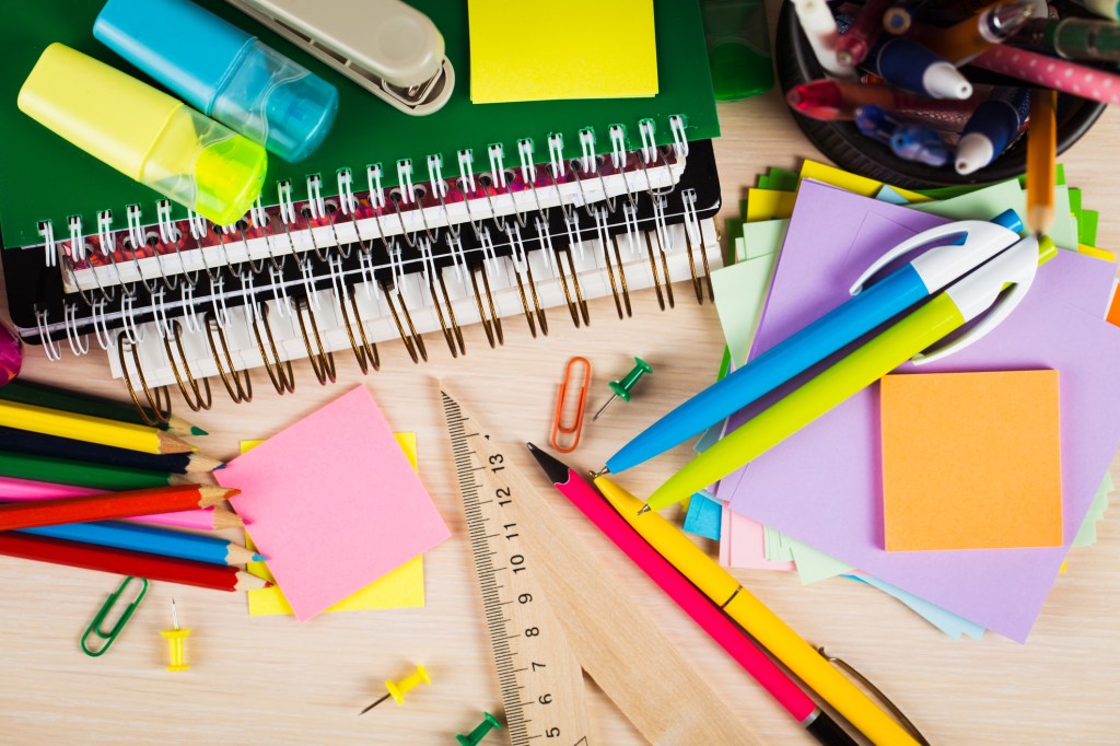 Materiais escolares e de escritório bem coloridos espalhados sobre uma mesa de madeira. Canetas, lápis, régua, post-it, cadernos, folhas...