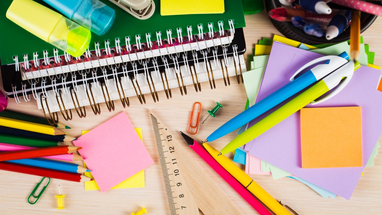 Materiais escolares e de escritório bem coloridos espalhados sobre uma mesa de madeira. Canetas, lápis, régua, post-it, cadernos, folhas...