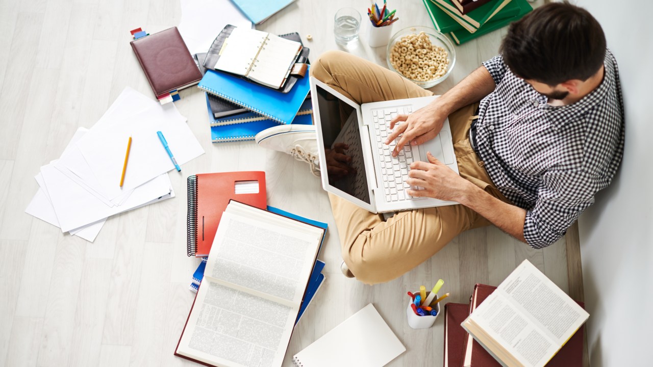 Jovem sentado no chão usando laptop, cercado por vários materiais universitários, como livros, cadernos, canetas e até um cereal