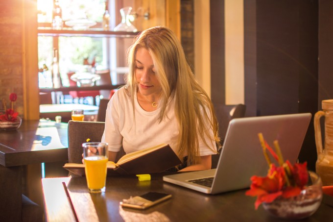 Garota estudando em um café, com um laptop e um livro, enquanto toma um suco