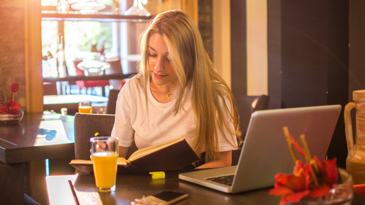 Garota estudando em um café, com um laptop e um livro, enquanto toma um suco