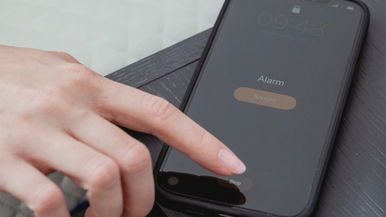 Fotografia foca em uma mão branca tocando na tela de um celular, que está tocando um alarme.