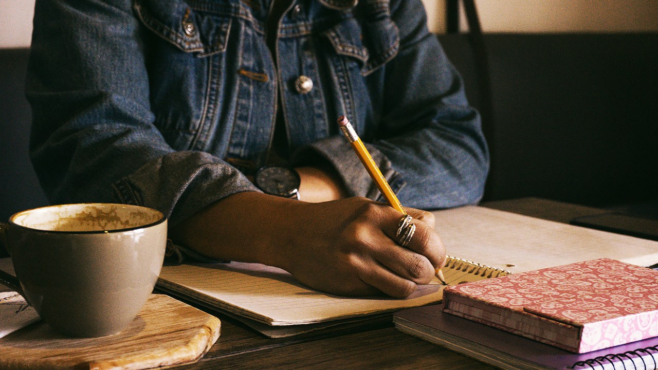 Estrutura da redação do Enem: saiba como desenvolver cada parte - Na imagem: uma mulher escrevendo sentada com uma caneca de café ao lado da mesa.