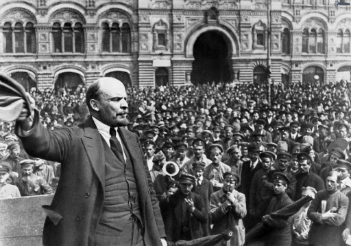 O líder Lenin, do partido bolchevique, acena para a população durante uma passeata. Nas ruas, uma multidão se aglomera para ver o político.