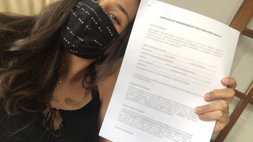 Segurando um contrato nas mãos, mulher tira uma selfie. Ela utiliza uma máscara de proteção e tem um semblante de comemoração.