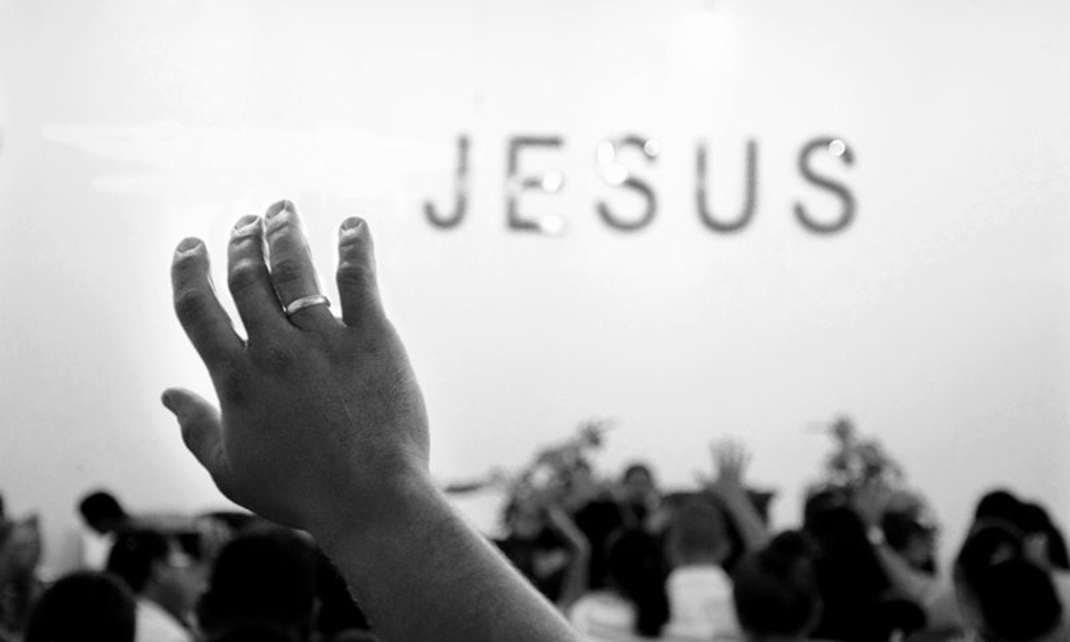 Mão levantada em sinal de oração. No fundo da imagem está escrito Jesus