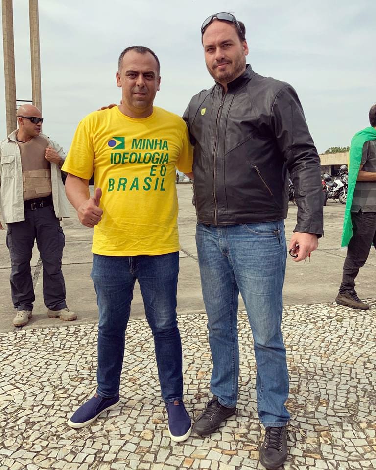 O deputado Anderson Moraes posa ao lado do vereador Carlos Bolsonaro (Republicanos) no dia 23 de maio de 2021. Ambos estão sem máscara. Moraes usa uma camisa amarela escrito em verde 