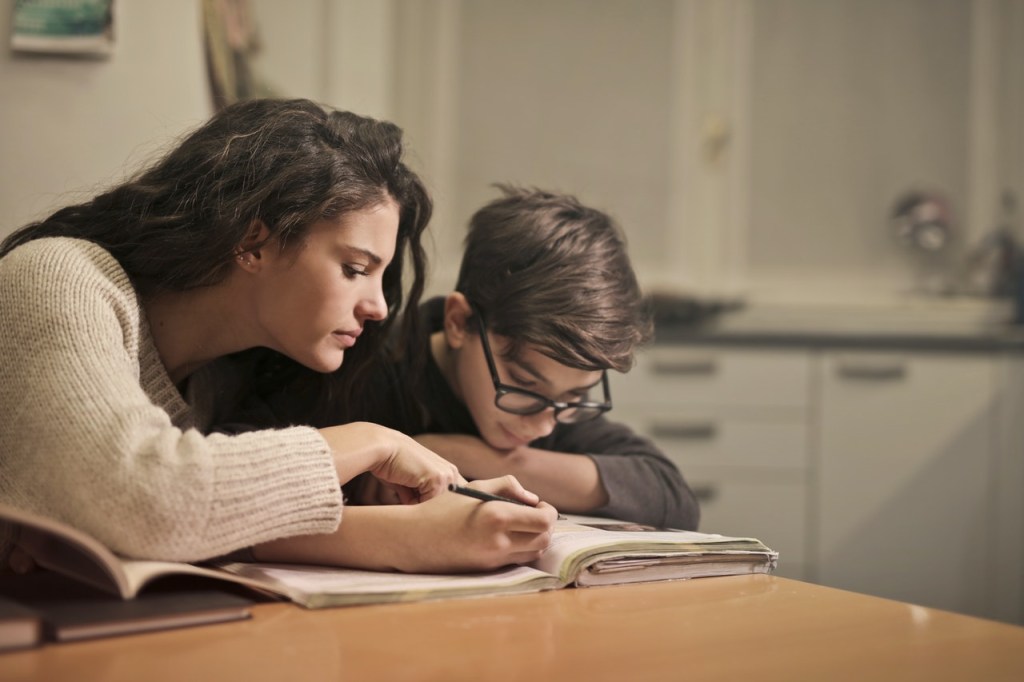 Mulher se apoia ao lado de um menino, que está lendo um livro. Os dois estão sentados em uma mesa dentro de um ambiente fechado.