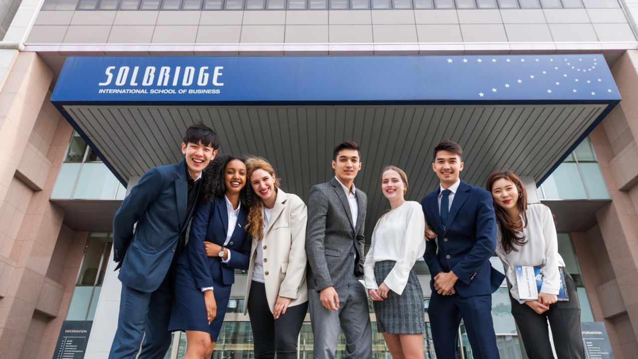 Grupo de 8 jovens, usando roupas empresariais, posa junto sorrindo em frente a um prédio da universidade, onde vemos o escrito "SolBridge: International School of Business"