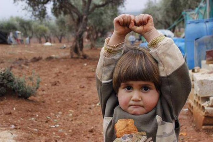 Adi Hudea, uma menina síria, ergue os braços e fecha as mãos em gesto de rendição, após confundir a câmera fotográfica com uma arma