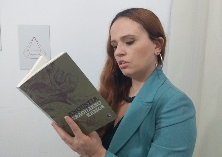 Mulher branca, de cabelos cobreados, usando um blazer turquesa, em pé, lendo o livro aberto 'Angústia', de Graciliano Ramos.