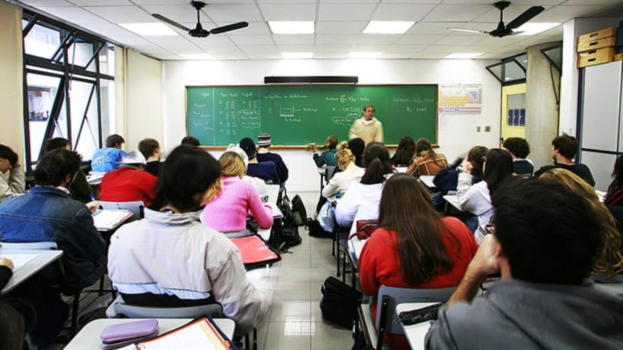 Foto de uma sala de aula, com uma professora dando aula enquanto alunos prestam atenção em suas carteiras.