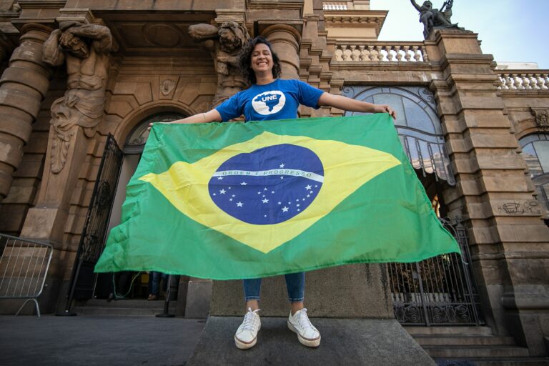 Jovem negra de cabelos castanhos na altura dos ombros veste camiseta azul com o símbolo da UNE enquanto estende a bandeira do Brasil com os dois braços. Ela está em frente ao Teatro Municipal, em São Paulo,