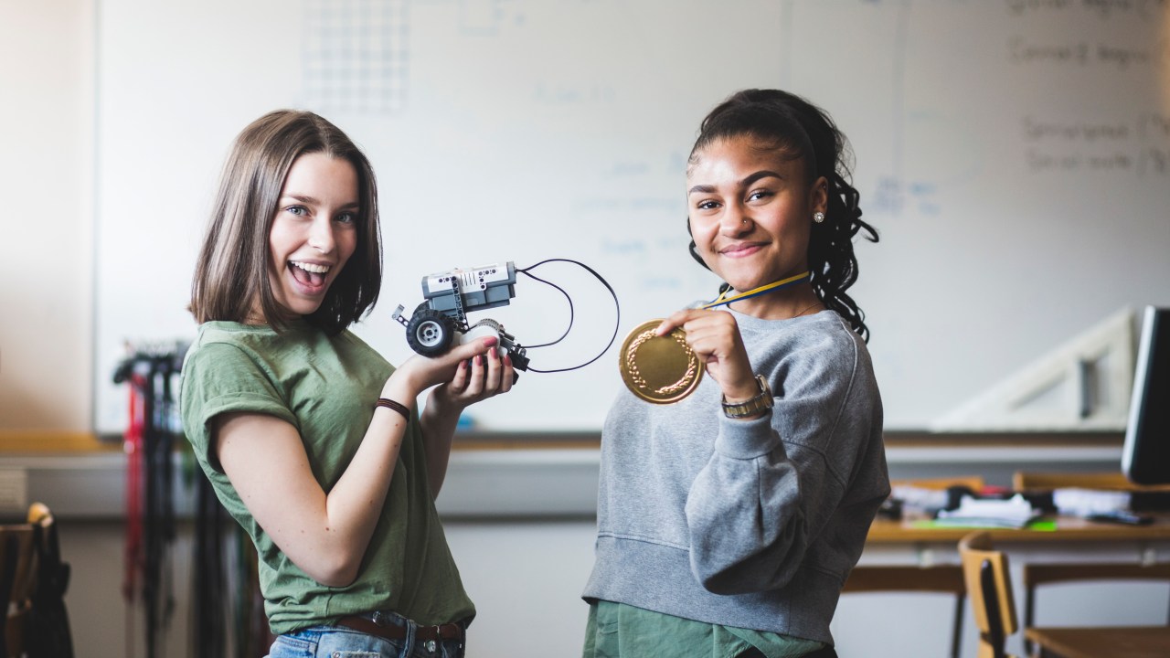 Alunas adolescentes bem-sucedidas segurando um robô e uma medalha de ouro em uma sala de aula no Ensino Médio