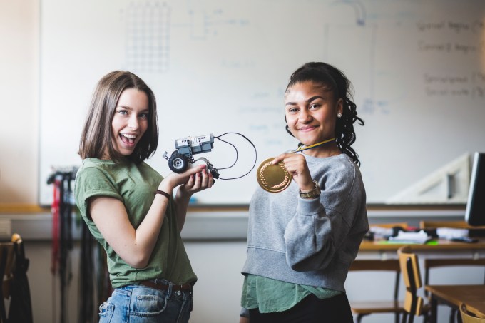 Portrait of successful female teenage students holding robot and gold medal while standing in classroom at high school