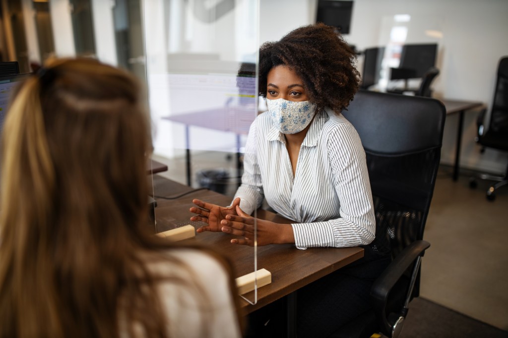 Mulheres conversam em um escritório. Elas usam máscaras e há um protetor de espirro entre elas.