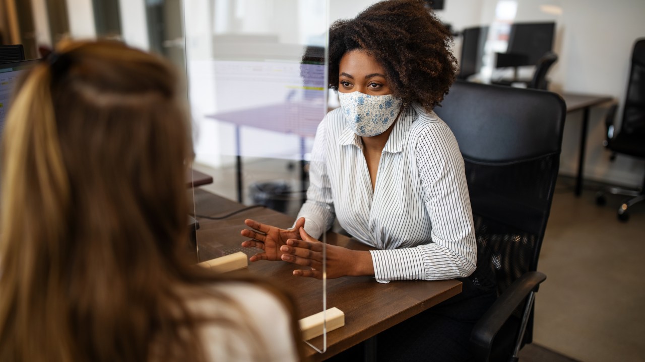 Mulheres conversam em um escritório. Elas usam máscaras e há um protetor de espirro entre elas.