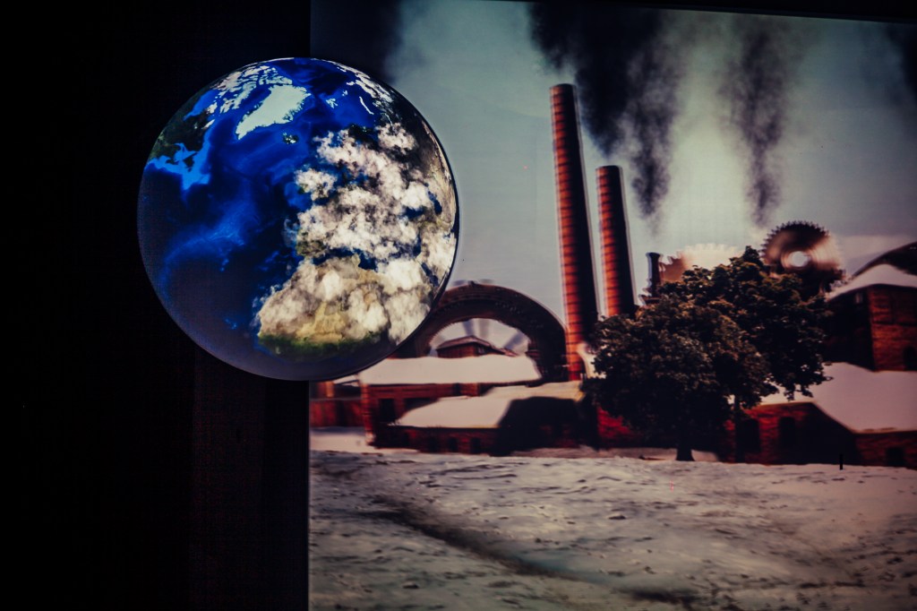 Paineis mostram o globo terrestre e, ao fundo, a imagem de uma fábrica, lançando fumaça aos ares.
