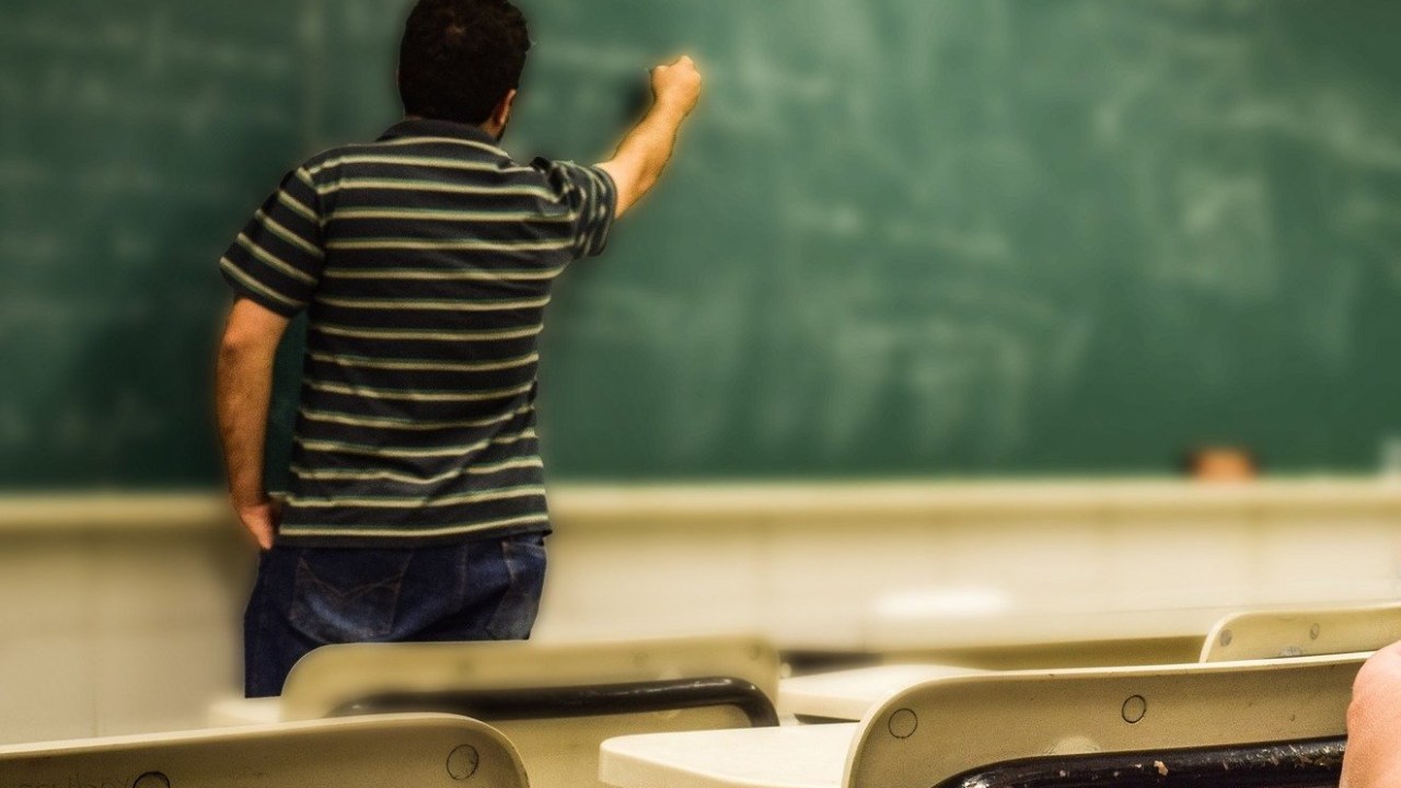 Professor de costas e usando camiseta listrada preta escreve em um quadro negro na sala de aula.