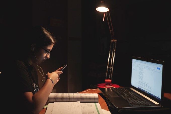 Student using smartphone while working with laptop at home in the dark