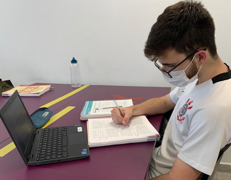 Estudante Luís Henrique Amaral Silva está em uma mesa com notebook, caderno e garrafa de água. Ele usa máscara e camiseta do Corinthians.
