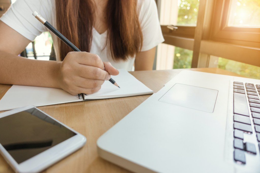 computador, celular e uma garota fazendo anotações em um papel