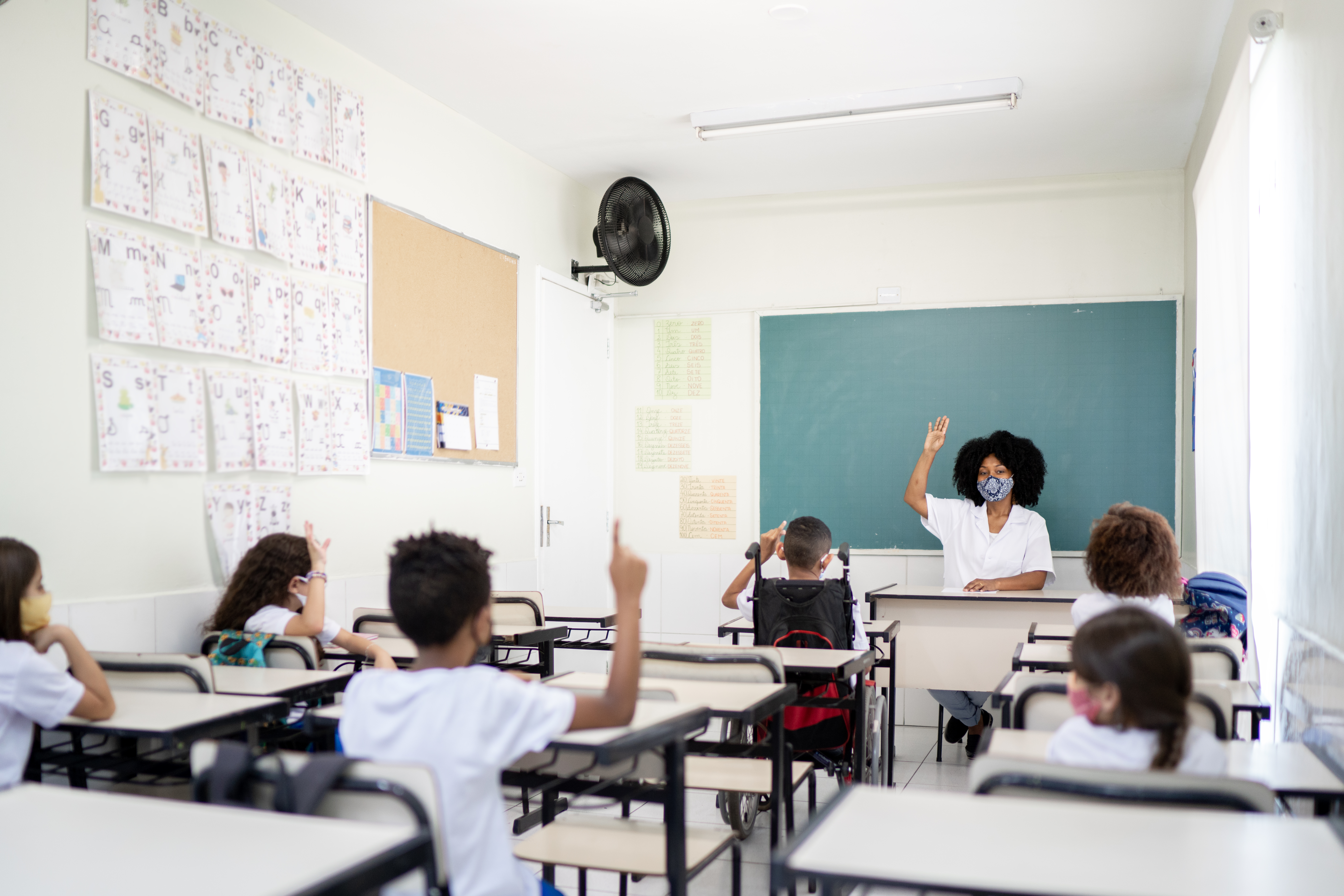 Sala de aula com uma professora negra e 6 alunos crianças. Todos estão de máscara e com distanciamento entre as carteiras.