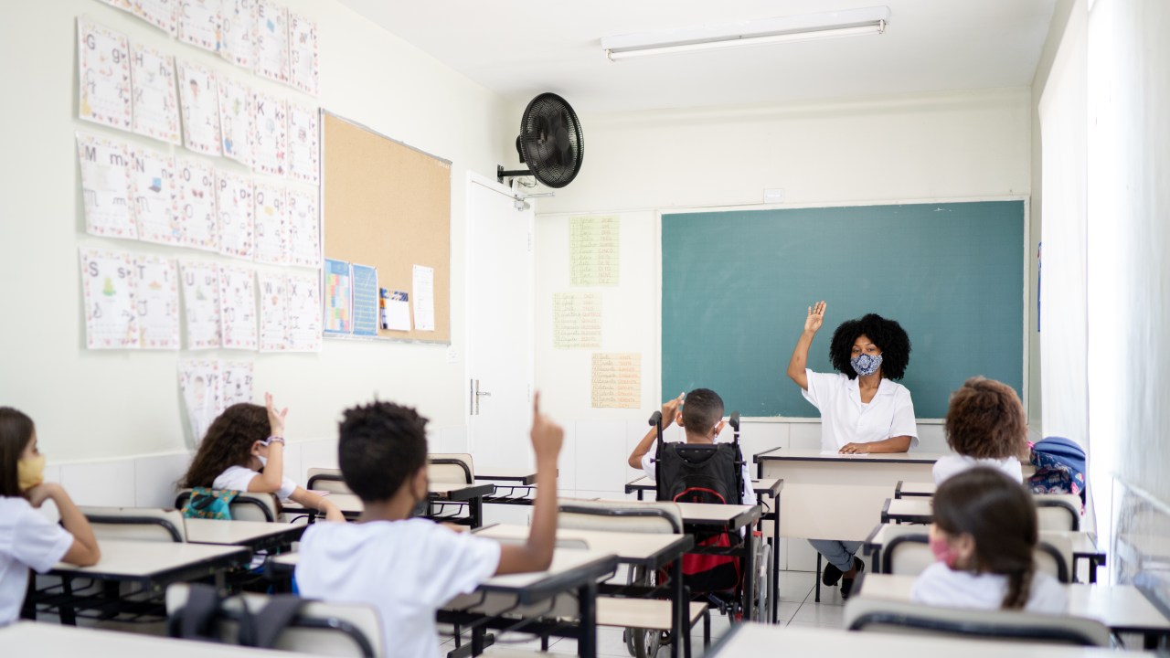 Sala de aula com uma professora negra e 6 alunos crianças. Todos estão de máscara e com distanciamento entre as carteiras.