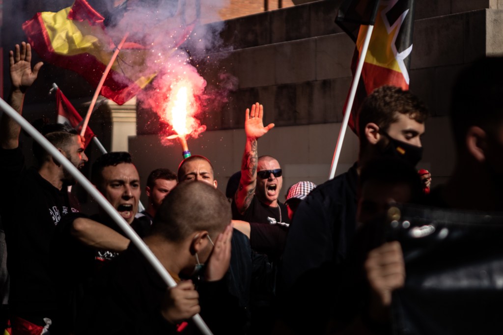 Fotografia mostra protesto com pessoas segurando bandeiras e um deles está levantando a mão em um gesto nazista
