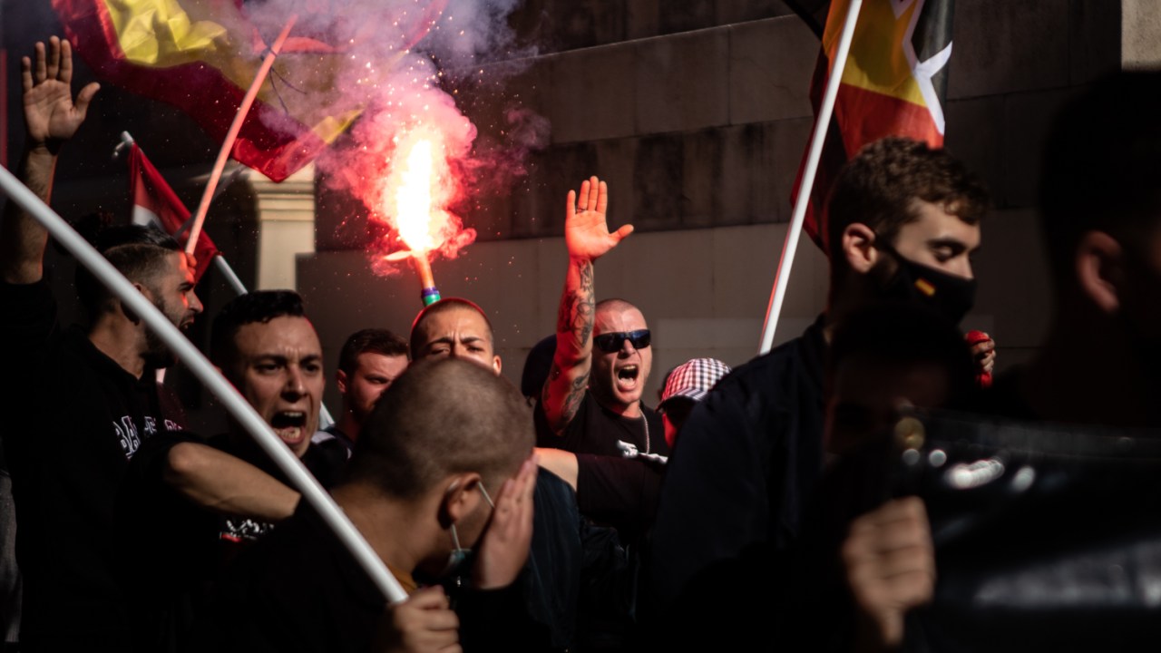 Fotografia mostra protesto com pessoas segurando bandeiras e um deles está levantando a mão em um gesto nazista