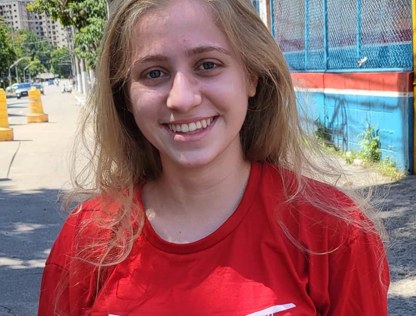 Mulher jovem branca, loira e de olhos claro, sorri para a câmera usando camiseta vermelha. Ela está na rua.
