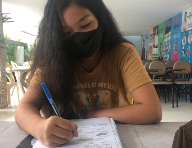 Jovem mulher usando máscara de proteção no rosto está sentada escrevendo em um livro. Ela tem pele morena e cabelos negros.