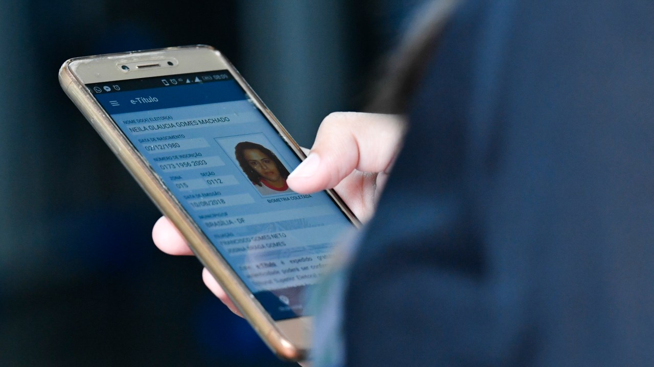 Foto de mão segurando celular com o aplicativo "e-Título" aberto. Há uma foto e informações sobre o usuário.