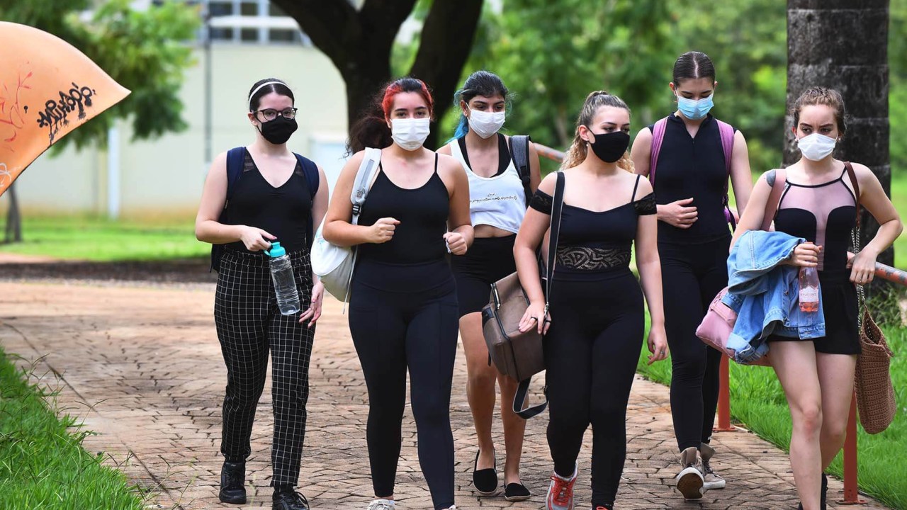 Grupo de jovens mulheres máscara. Algumas estão usando roupas esportivas, e outras estão com bolsas, casacos e garrafa d'água na mão