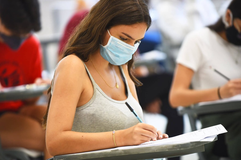 Jovem mulher usando máscara está sentada escrevendo de caneta em sua prova do vestibular. Ela está em uma sala de aula de aplicação da prova