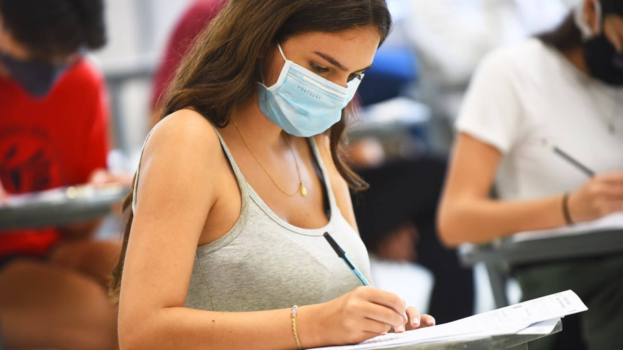 Jovem mulher usando máscara está sentada escrevendo de caneta em sua prova do vestibular. Ela está em uma sala de aula de aplicação da prova