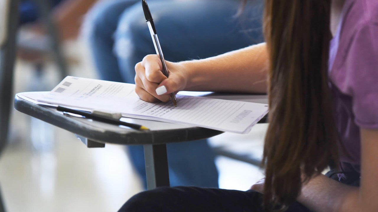 Mão de uma jovem mulher sentada escrevendo de caneta em sua prova de redação do vestibular. Ela está em uma sala de aula de aplicação da prova