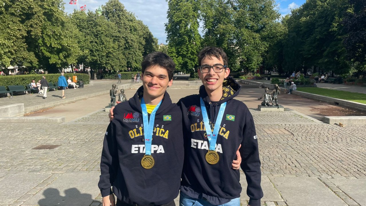 Dois rapazes com medalhes de ouro no pescoço posam com bandeira do Brasil em uma praça públcia ensolarada