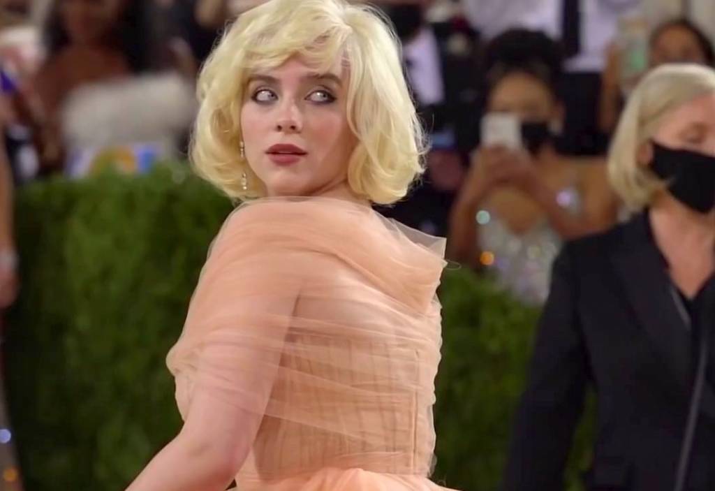 Billie Eilish A cantora Billie Eilish tem pele branca clara, cabelos loiros acima dos ombros, olhos azuis e usa um vestido salmão.