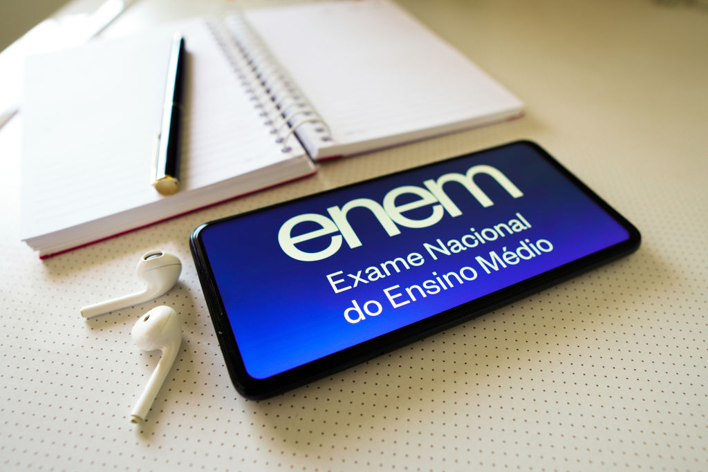 Celular com a tela escrito "Enem", ao lado de um caderno e dois fones de ouvido.