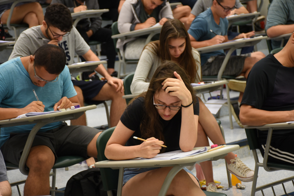 Fotografia foca em parte de uma sala de aula de aplicação de prova. O foco principal é uma menina concentrada em sua prova, ela está com a mão na cabeça e com um lápis na outra mão.